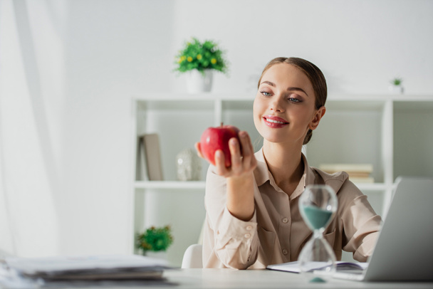 щаслива бізнес-леді тримає яблуко на робочому місці з піщаним годинником і ноутбуком в офісі
  - Фото, зображення