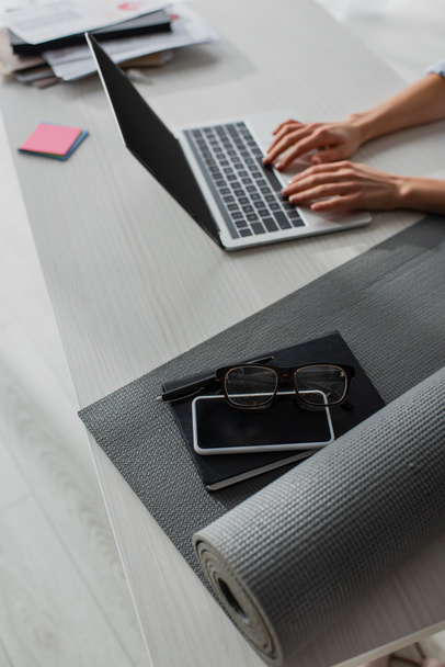 cropped view of businesswoman working on laptop at workplace with fitness mat, notepad, smartphone and eyeglasses - Photo, Image