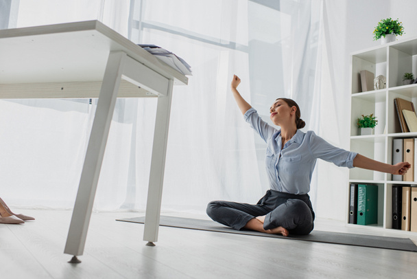 fiatal üzletasszony nyújtva kezét és gyakorló jóga lótuszpozícióban matracon az irodában  - Fotó, kép