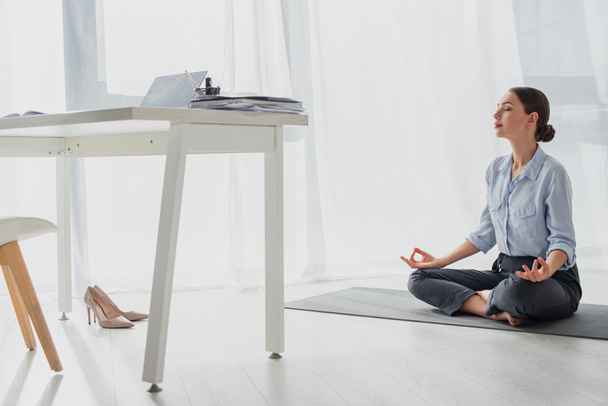 mladá podnikatelka cvičit jógu v lotosové pozici s gyan mudra na podložce v kanceláři  - Fotografie, Obrázek