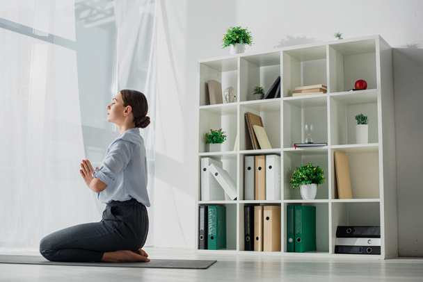 Geschäftsfrau praktiziert Yoga und meditiert mit namaste Geste auf Matte im Büro  - Foto, Bild