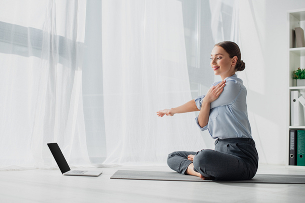 młoda przedsiębiorczyni mająca zajęcia online na laptopie i wyciągająca ręce w pozycji lotosu na macie w biurze  - Zdjęcie, obraz