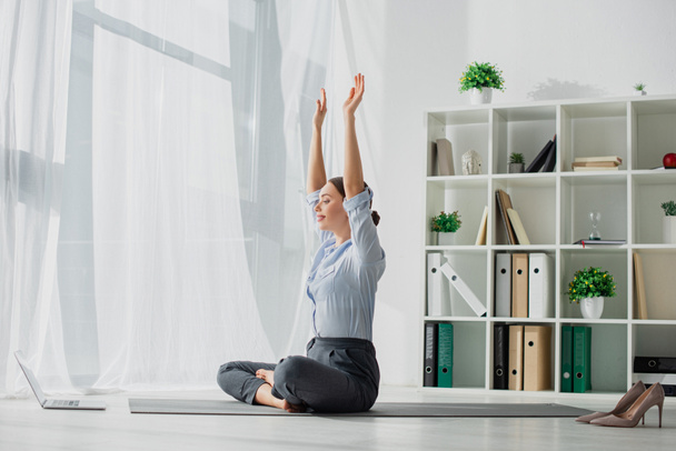 jovem empresária ter aulas on-line sobre laptop e praticar ioga na posição de lótus no tapete com as mãos para cima no escritório
  - Foto, Imagem