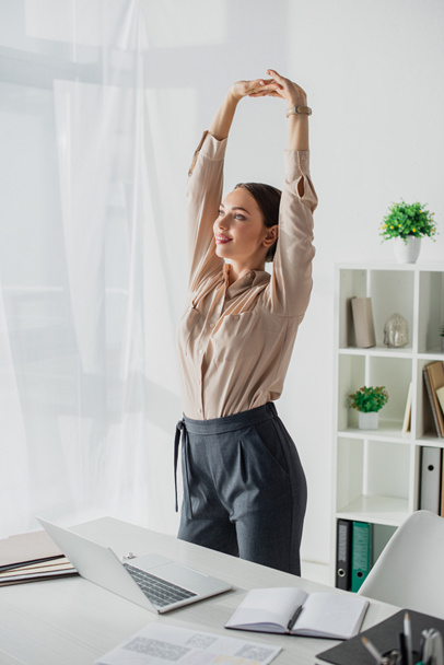 krásný mladý businesswoman stretching arms v moderní kanceláři s notebookem - Fotografie, Obrázek