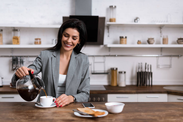 smiling businesswoman pouring coffee in cup near smartphone with blank screen while hiding problem of domestic violence  - Фото, зображення