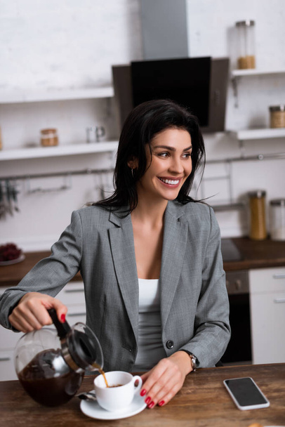 smiling businesswoman pouring coffee in cup near smartphone with blank screen while hiding problem of domestic violence  - Photo, Image