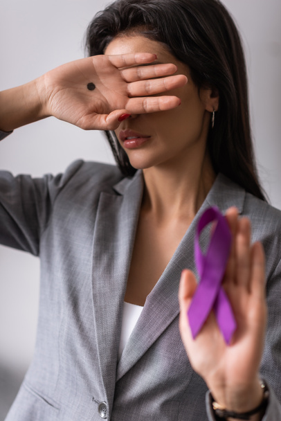вибірковий фокус ділової жінки з чорною крапкою на долоні, що закриває обличчя та тримає фіолетову стрічку на сірій концепції домашнього насильства
  - Фото, зображення