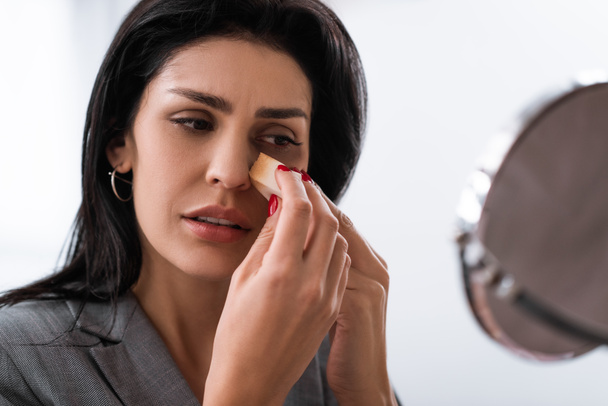 selective focus of sad woman with bruise on face holding makeup sponge while applying makeup foundation near mirror, domestic violence concept  - Photo, Image