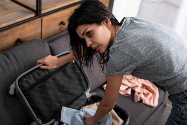 selective focus of sad woman with bruise on face packing suitcase with clothing, domestic violence concept  - Foto, Imagen