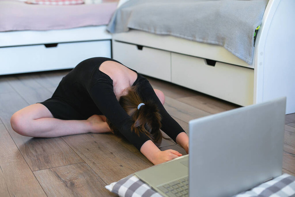 Schoolgirl watching online video on laptop and doing sports exercises - yoga, gymnastics, choreography. Stay at home. Concept of self isolation, quarantine, online education. - Fotó, kép