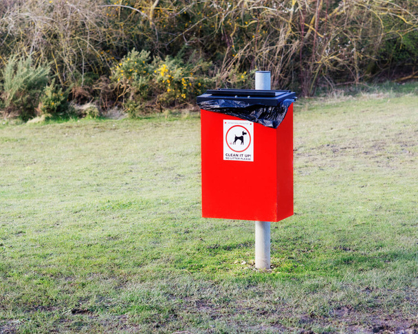 Κόκκινος σκουπιδοτενεκές για τα σκουπίδια του σκύλου με πινακίδα καθαρίστε το και τη φωτογραφία ενός σκύλου στο πάρκο. - Φωτογραφία, εικόνα