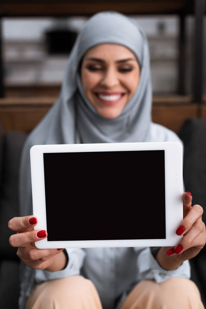 selective focus of positive arabian woman in hijab holding digital tablet with blank screen in living room  - Фото, зображення