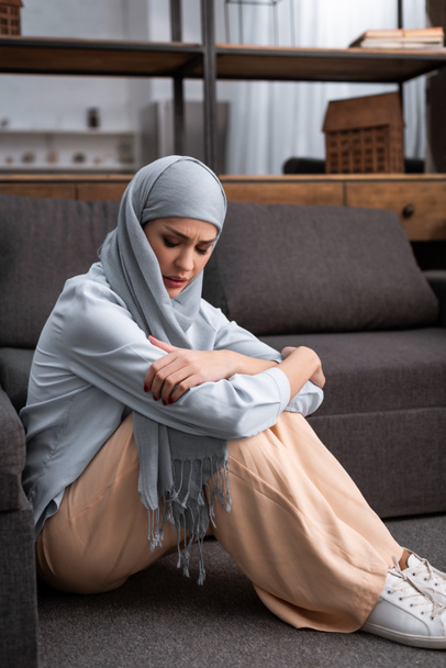 αναστατωμένη αραβίδα γυναίκα με μαντίλα κάθεται στο πάτωμα στο σαλόνι, έννοια της ενδοοικογενειακής βίας   - Φωτογραφία, εικόνα