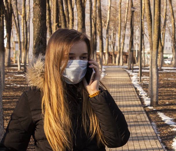 A girl in a protective mask walks in the Park. A woman in a medical mask is talking on the phone. The epidemic of coronavirus. Protection from Covid-19. - Foto, Bild