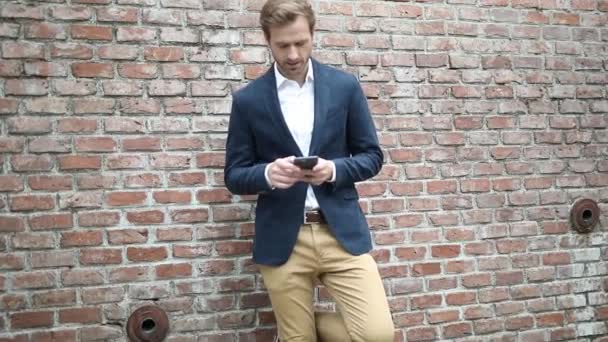 Όμορφος ξανθός επιχειρηματίας που φοράει ναυτικό κοστούμι γράφει ένα μήνυμα στο κινητό του ενώ γέρνει σε έναν τοίχο από τούβλα και δείχνει προς τα εμπρός - Πλάνα, βίντεο