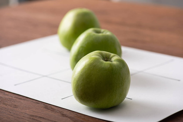 selective focus of tic tac toe game with row of three green apples on white paper on wooden surface - Photo, Image