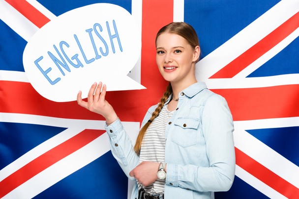χαμογελαστό όμορφο κορίτσι με πλεξούδα κρατώντας φούσκα ομιλία με τα αγγλικά γράμματα στο φόντο σημαία Ηνωμένο Βασίλειο - Φωτογραφία, εικόνα
