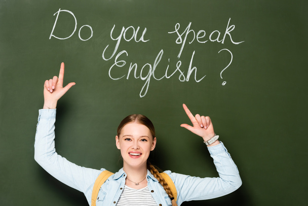 smiling girl with backpack pointing at chalkboard with do you speak English lettering - Foto, imagen