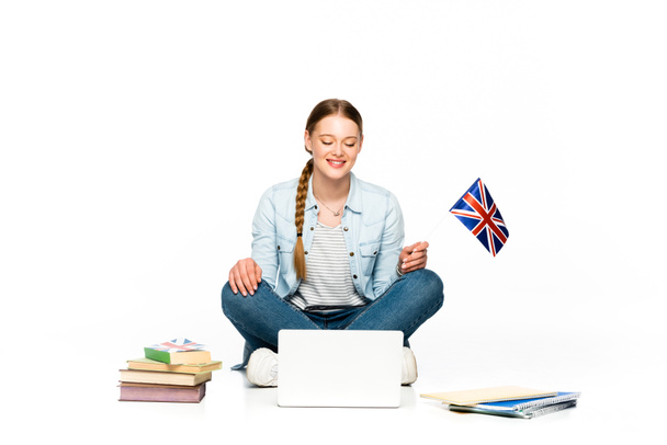 χαμογελαστό κορίτσι κάθεται στο πάτωμα με φορητό υπολογιστή, βιβλία και βιβλία και κρατώντας σημαία Ηνωμένο Βασίλειο απομονώνονται σε λευκό - Φωτογραφία, εικόνα