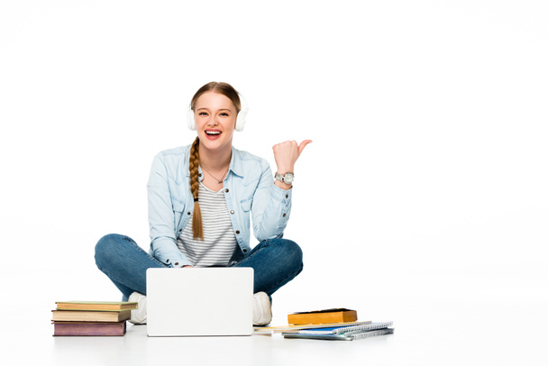 smiling girl sitting on floor in headphones near laptop, books and copybooks and pointing aside isolated on white - Photo, image