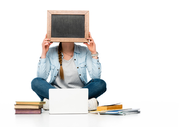 girl sitting on floor with obscure face near laptop, books and copybooks and holding chalkboard isolated on white - Фото, изображение