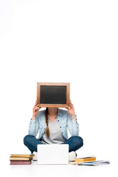 girl sitting on floor with obscure face near laptop, books and copybooks and holding chalkboard isolated on white - Foto, Imagem
