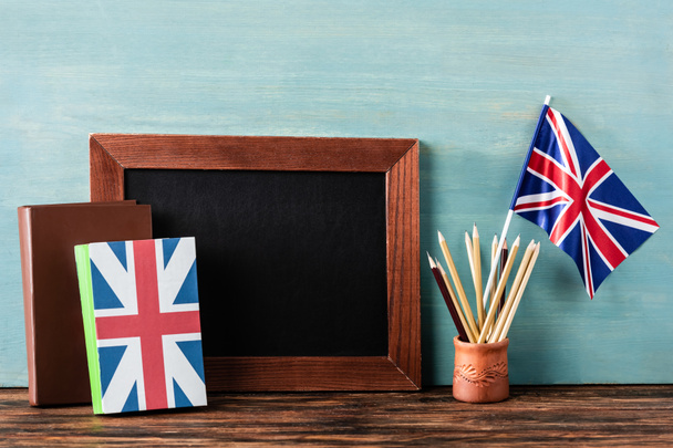 empty chalkboard near pencils, books and uk flag on wooden table near blue wall - Photo, Image