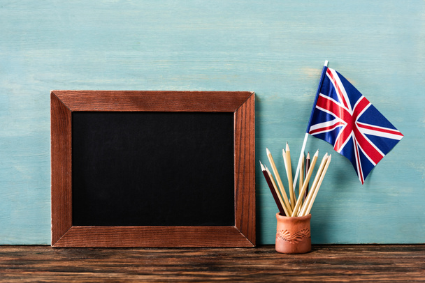 empty chalkboard near pencils and uk flag on wooden table near blue wall - Фото, изображение