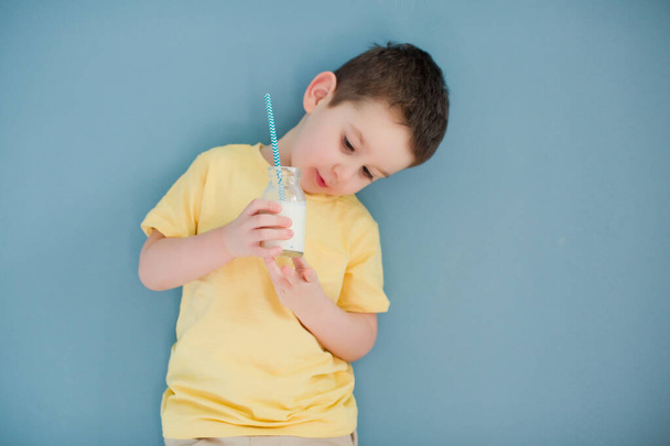 Niño caucásico en una camiseta amarilla beber leche de botella de vidrio y paja
 - Foto, Imagen