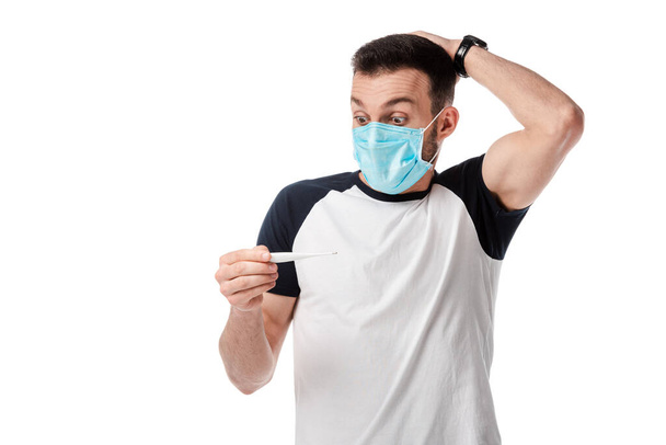 σοκαρισμένος άνθρωπος με ιατρική μάσκα κοιτάζοντας ψηφιακό θερμόμετρο απομονωμένο σε λευκό  - Φωτογραφία, εικόνα
