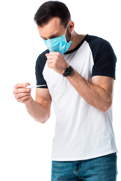άνθρωπος με ιατρική μάσκα κοιτάζοντας ψηφιακό θερμόμετρο και βήχα απομονώνονται σε λευκό  - Φωτογραφία, εικόνα