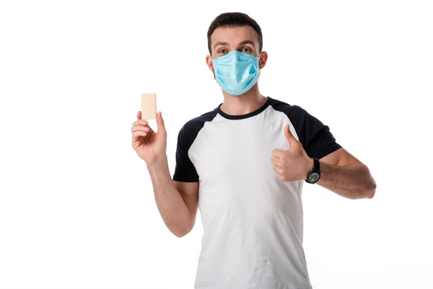 άνθρωπος με ιατρική μάσκα δείχνει τον αντίχειρα και κρατώντας σαπουνόπερα απομονωμένη σε λευκό  - Φωτογραφία, εικόνα
