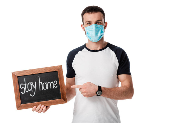 man in medical mask pointing with finger at chalk board with stay home lettering isolated on white  - Zdjęcie, obraz