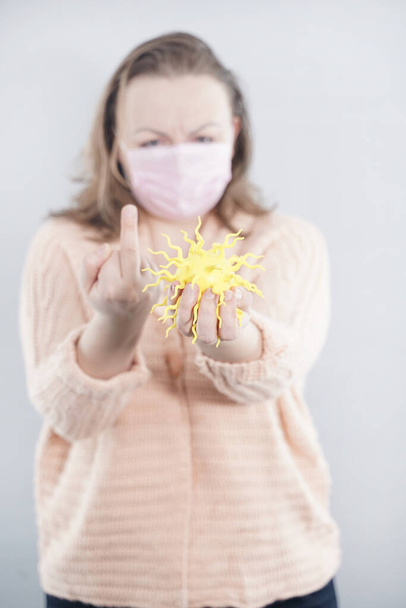 μια γυναίκα με προστατευτική μάσκα κρατά στο χέρι της ένα βακτήριο coronavirus και εκφράζει αρνητικά συναισθήματα για την επιδημία στο γκρίζο φόντο - Φωτογραφία, εικόνα