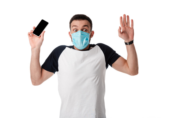 shocked man in medical mask holding smartphone with blank screen and putting hands up isolated on white  - Foto, afbeelding