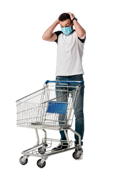 shocked man in medical mask looking at empty shopping cart isolated on white - Foto, imagen