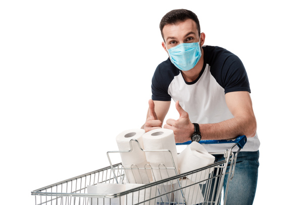 Tıbbi maskeli bir adam, beyaz üzerine izole edilmiş tuvalet kağıtlarıyla alışveriş arabasının yanında baş parmağını gösteriyor. - Fotoğraf, Görsel