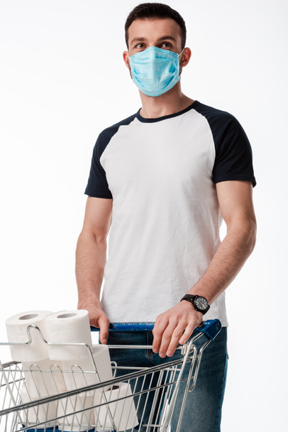 muž v lékařské masce stojící poblíž nákupního košíku s toaletním papírem izolovaným na bílém - Fotografie, Obrázek