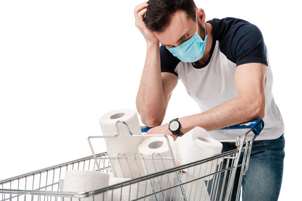 muž v modré lékařské masce při pohledu na nákupní košík s toaletním papírem izolovaným na bílém - Fotografie, Obrázek