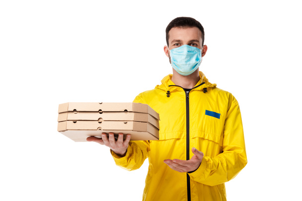 dostawca w masce medycznej wskazując ręką na pudełka pizzy odizolowane na białym  - Zdjęcie, obraz