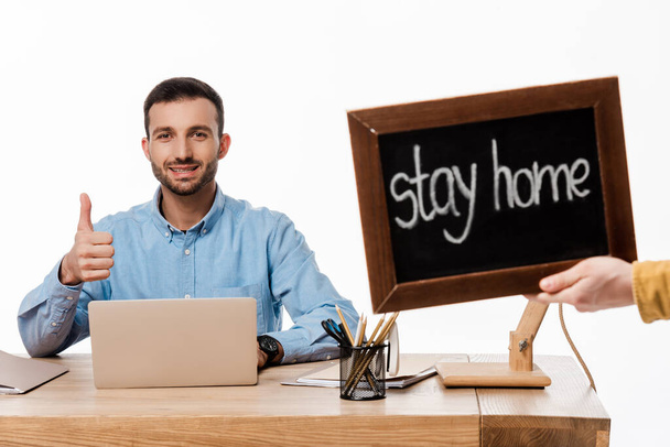 happy freelancer showing thumb up near laptop and chalkboard with stay home lettering isolated on white  - Foto, Imagen