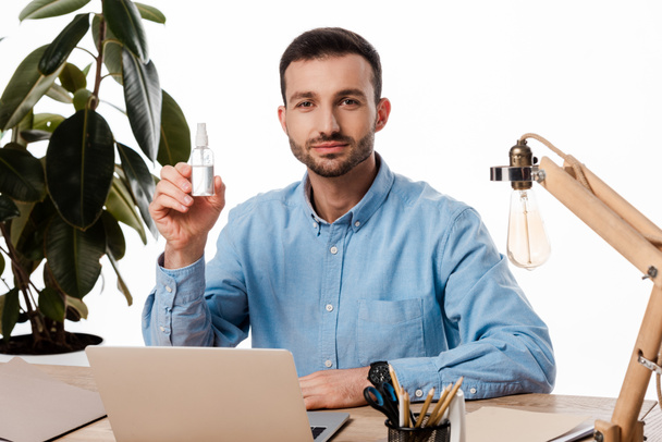 handsome freelancer holding hand sanitizer near laptop isolated on white  - Фото, изображение