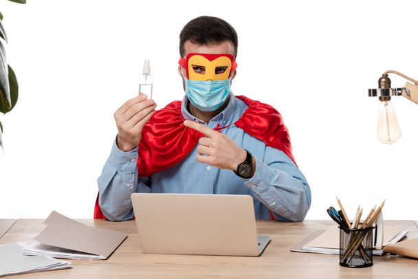 muž v lékařské masce a superhrdina kostým ukazuje prstem na ruce dezinfekci v blízkosti notebooku izolované na bílé  - Fotografie, Obrázek