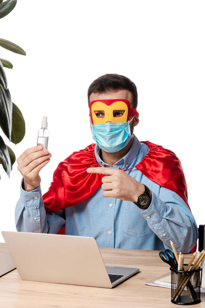 Mann in medizinischer Maske und Superheldenmantel zeigt mit dem Finger auf Handdesinfektionsmittel in der Nähe Laptop isoliert auf weiß  - Foto, Bild