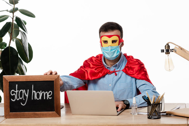 freelancer em máscara médica e traje de super-herói segurando placa de giz com ficar em casa lettering isolado no branco
  - Foto, Imagem