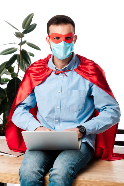 freelancer in medical mask and superhero costume using laptop and sitting on table isolated on white  - 写真・画像