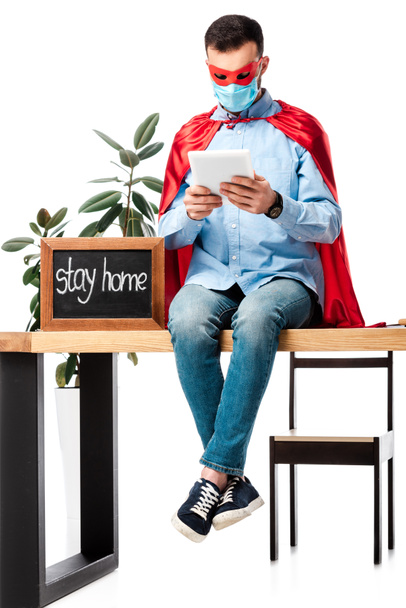 man in medical mask and superhero cape using digital tablet near chalk board with stay home lettering isolated on white  - Foto, imagen