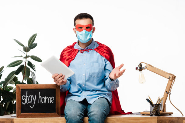 άνθρωπος με ιατρική μάσκα και κάπα υπερήρωα κρατώντας ψηφιακή ταμπλέτα κοντά στον πίνακα κιμωλία με παραμονή στο σπίτι γράμματα απομονώνονται σε λευκό  - Φωτογραφία, εικόνα