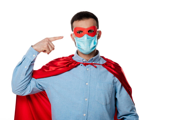 Mann im Superheldenkostüm mit Umhang zeigt mit dem Finger auf medizinische Maske isoliert auf weiß   - Foto, Bild