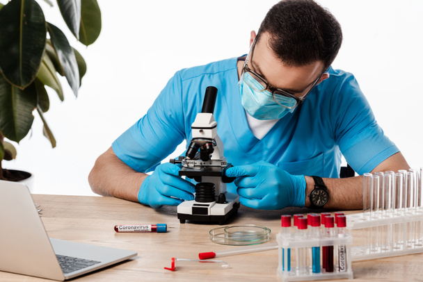 Wissenschaftler mit medizinischer Maske und Brille, Mikroskop in der Nähe von Reagenzgläsern mit Coronavirus-Schriftzug und Laptop isoliert auf weiß  - Foto, Bild